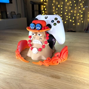 Peut inclure: Un support de manette Xbox à thème de personnage, de couleur orange et beige, avec une manette Xbox blanche posée dessus. Le support présente un chapeau rouge, des lunettes bleues et un collier de perles rouges. Sur une surface en bois.