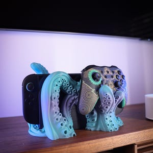 Könnte beinhalten: Ein Gaming-Controller- und Konsolenständer im Tintenfisch-Design in Türkis und Lila. Der Ständer ist so konzipiert, dass er eine Spielekonsole und einen Controller aufnehmen kann, wobei sich die Tentakel um die Geräte wickeln. Der Controller hat ein schwarzes, türkisfarbenes und goldenes Design.