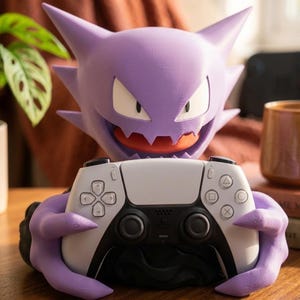 Puede incluir: Una figura de personaje morado con cabeza puntiaguda, ojos blancos y una amplia sonrisa con dientes, sosteniendo un mando de videojuego blanco y negro. El personaje tiene orejas puntiagudas y manos en forma de garra que sujetan el mando.