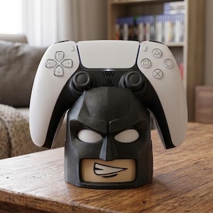 Puede incluir: Un soporte para mando con temática de Batman en color negro, con forma de máscara con ojos blancos y boca, que sostiene un mando de juego inalámbrico blanco y negro. El soporte presenta una superficie texturizada y un gráfico estilizado.