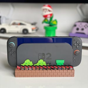 Può includere: Una console Nintendo Switch grigia inserita in un supporto in plastica marrone con motivo a mattoni. Il supporto presenta cespugli verdi pixelati e un tubo verde, con fessure che contengono diverse cartucce di gioco.