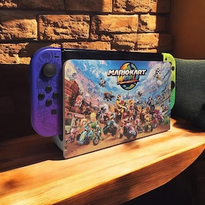 May include: A Nintendo Switch console with a purple Joy-Con controller on the left and a green Joy-Con on the right. The screen displays a colorful graphic from the Mario Kart World game, featuring various characters and vehicles.