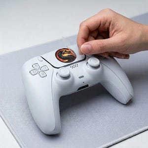 Könnte beinhalten: Ein weißer Videospiel-Controller mit einem Aufkleber mit einem Drachenlogo, der angebracht wird. Der Controller hat ein schlankes Design mit Tasten und Joysticks. Der Aufkleber ist rund und hat rote, gelbe und schwarze Farben.