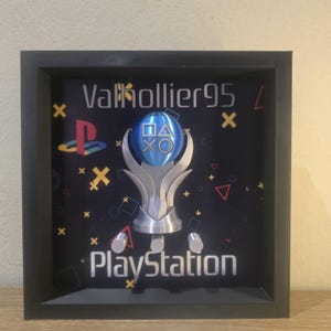 Puede incluir: Un marco cuadrado negro tipo caja con una escultura de trofeo plateada que tiene una esfera azul con símbolos de mando de PlayStation. El fondo es negro con el texto Valkollier95 y PlayStation, además de formas geométricas coloridas.