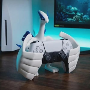 May include: A white and blue Lugia figurine acting as a controller stand, holding a white and black wireless game controller. The figure features a long neck, pointed head, and large wing-like hands that securely grip the gaming peripheral.