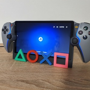 May include: A handheld gaming device with a screen showing a blue interface and the PlayStation logo. It has grey controllers with blue lighting. Colourful triangle, circle, X, and square buttons are in front.