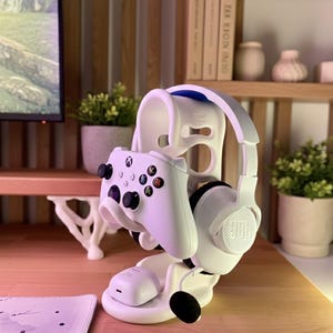 May include: A white gaming headset and Xbox controller are displayed on a white stand. The stand includes a charging base and sits on a wooden surface. The headset features a JBL logo.