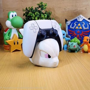 May include: A white and black PlayStation 5 controller sits on a white Mewtwo-shaped stand with a pink eye. Other figurines of video game characters are in the background, including Yoshi, a star, and Pokemon characters.