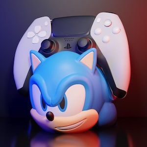 Puede incluir: Una figura azul de Sonic the Hedgehog con un mando de PlayStation 5 blanco y negro encima. La figura tiene una expresión sonriente y está sobre un fondo degradado azul y rojo.