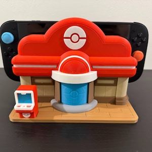 Peut inclure: Modèle imprimé en 3D rouge et blanc d'un Centre Pokémon, avec une petite borne d'arcade sur une base marron. Une Nintendo Switch noire est derrière le modèle. Le modèle a un toit rouge avec un symbole Poké Ball blanc.