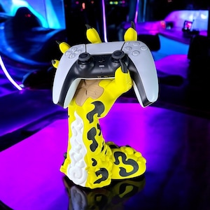 May include: A white and black game controller rests on a yellow and black claw-shaped stand. The stand has a unique design with a textured pattern and yellow claw-like extensions. The controller is a modern design, and the stand is on a reflective surface.