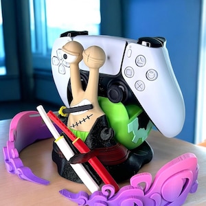 May include: A 3D printed snail-shaped controller stand featuring a tan body with a stitched mouth, holding a black sword in its mouth. It has a black base, a green shell, and two miniature swords in red and white. A white game controller rests on it.