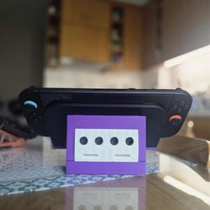 May include: A black handheld gaming console is mounted on a purple and white stand. The stand has a rectangular base with four circular holes and two horizontal slots. The console has blue and orange buttons. The stand is on a glass table.