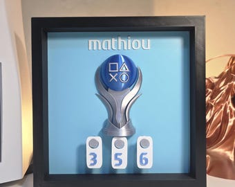 Puede incluir: Un marco cuadrado negro tipo caja con fondo azul claro y el nombre "mathiou" en letras blancas. Centrado hay un trofeo plateado con una esfera azul que muestra los símbolos de PlayStation en blanco. Debajo, tres fichas blancas con los números 3, 5, 6.