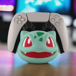 May include: A light gray PlayStation 5 controller rests on a teal Bulbasaur-shaped stand. The stand has red eyes, a red mouth, and green accents. The controller is white with black joysticks.