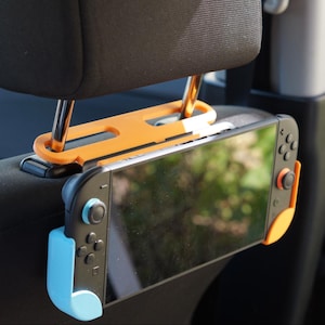 Può includere: Un supporto arancione e nero per poggiatesta per auto che tiene una console di gioco Nintendo Switch. Il supporto è progettato per essere fissato ai montanti del poggiatesta, offrendo un'esperienza di gioco sicura per i passeggeri. Lo Switch ha controller blu e arancioni.
