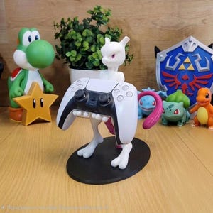 Può includere: Una statuetta bianca di Mewtwo su una base ovale nera, progettata per sostenere un controller di gioco bianco e nero. Il personaggio ha occhi viola e una lunga coda rosa, posizionato davanti a una pianta verde e ad altre figure.
