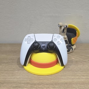 Puede incluir: Un mando de PlayStation 5 blanco y negro apoyado en un soporte con forma de sombrero amarillo y rojo. Una figura de caracol de dibujos animados está en el fondo. El mando tiene un diseño elegante con detalles en negro.