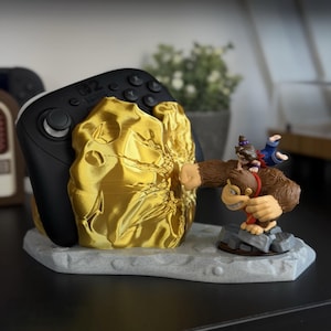 May include: A black Nintendo Switch controller rests in a gold-colored, textured holder. A figurine of Donkey Kong and Diddy Kong is on the base. The base is gray and has a textured surface. The controller is black with gray buttons.