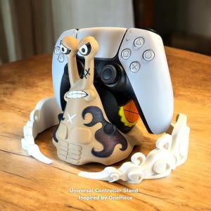 以下が含まれることがあります： 白と黒のゲームコントローラーを保持する、漫画のカタツムリのデザインが特徴のベージュ色のコントローラー スタンド。スタンドには雲のような装飾的なベースがあり、「Universal Controller Stand - Inspired by OnePiece」というテキストがあります。