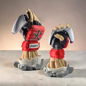 Può includere: Due supporti a forma di mano scolpita per controller di gioco, ciascuno con un guanto da arti marziali nero e rosso con la scritta FIGHT e un carattere giapponese. Le mani emergono da una base in pietra crepata, una sostiene un controller bianco e nero.