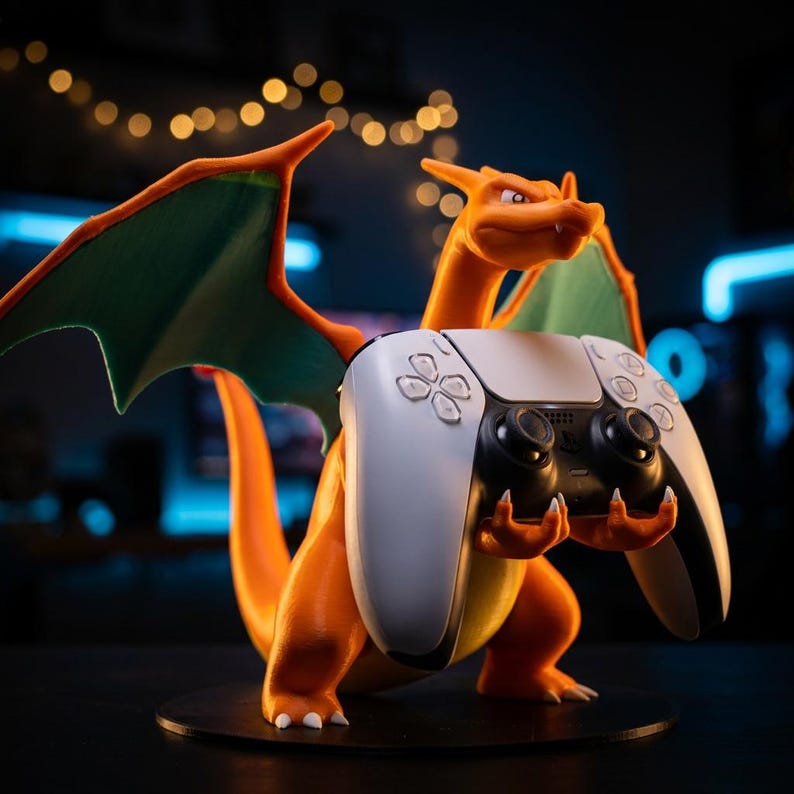 Puede incluir: Una figura naranja y verde con forma de drag&oacute;n sobre una base circular, que sostiene un mando de videojuego inal&aacute;mbrico blanco y negro en sus manos. La figura presenta grandes alas, una cola larga y una cabeza detallada con peque&ntilde;os dientes blancos.