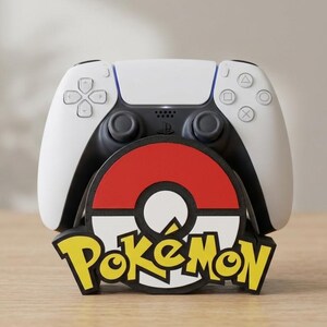 May include: A white and black video game controller resting on a circular stand featuring a red, white, and black Poké Ball design with the yellow and black Pokémon logo text displayed prominently across the front of the base.