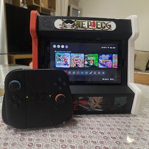 Könnte beinhalten: Ein schwarzer Nintendo Switch-Controller neben einer rot-schwarz-weißen Spielkonsole im Arcade-Stil. Die Konsole zeigt mehrere Videospielsymbole an. Auf der Oberseite der Konsole steht der Text "ONE PIECE".