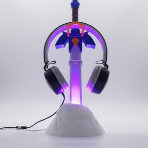 Può includere: Un supporto per cuffie da gioco a forma di spada, con una luce viola. Il supporto è blu, arancione e bianco. Le cuffie sono nere e grigie. Il supporto è su una base bianca.