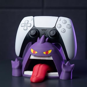 Peut inclure: Un support de manette en forme de Gengar violet, doté d'une bouche grande ouverte avec une langue rouge saillante, des yeux jaunes à pupilles rouges et deux petites mains. Une manette de jeu sans fil blanche et noire repose sur la figurine.