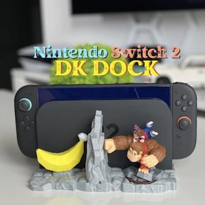 May include: A black gaming console dock featuring a grey rocky base with a yellow banana and a Donkey Kong figure. The text Nintendo Switch 2 DK DOCK is displayed above the console. The dock has black controllers attached to each side.