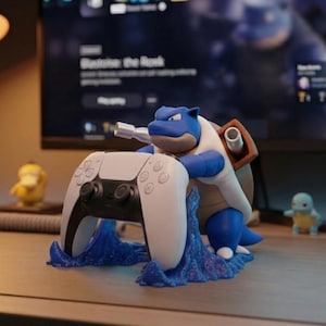 Könnte beinhalten: Eine blau-weiße Blastoise-Figur hält einen weißen PS5-Controller. Die Figur steht auf einem blauen Sockel, mit anderen Pokémon-Figuren im Hintergrund. Eine Schreibtischlampe und ein Monitor mit einem Spiel sind ebenfalls zu sehen.