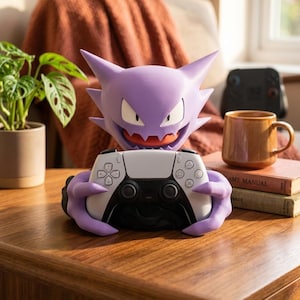 May include: A purple Gengar character figure with pointed ears and a wide grin, designed as a controller stand. The figure is holding a white and black wireless game controller with its hands. The surface of the figure is smooth and matte.