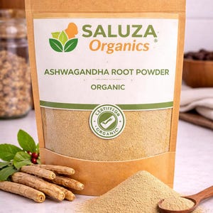 Puede incluir: Bolsa de papel marr&oacute;n con polvo de ra&iacute;z de ashwagandha org&aacute;nico, con la marca "SALUZA Organics". Un peque&ntilde;o mont&oacute;n de polvo y una cuchara de madera est&aacute;n frente a la bolsa. Tambi&eacute;n se ven varias ra&iacute;ces de ashwagandha.