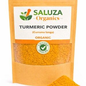 Puede incluir: Un paquete de cúrcuma en polvo con la marca "SALUZA - Organics" y el texto "TURMERIC POWDER (Curruma longa) ORGANIC". Un pequeño montón de polvo amarillo está al lado del paquete. El paquete tiene una ventana transparente.