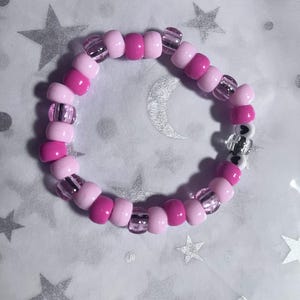 Könnte beinhalten: Ein Armband aus rosa und magenta Perlen, mit transparenten, zylindrischen Perlen dazwischen. Das Armband liegt auf einem weißen Hintergrund mit silbernen Sternen und einem Mond.