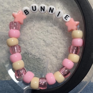 May include: A beaded bracelet with the name "BUNNIE" spelled out in white letter beads. The bracelet features pink, yellow, and clear beads, with pink star-shaped beads on each side of the name.