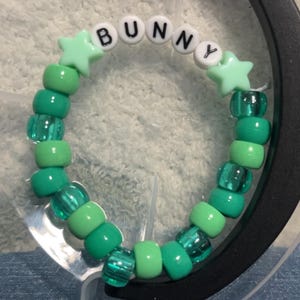 Könnte beinhalten: Ein grünes Perlenarmband mit dem Wort "BUNNY" in weißen Buchstabenperlen. Das Armband hat verschiedene Grüntöne und sternförmige Akzente. Das Armband befindet sich in einem transparenten Kunststoffbehälter.