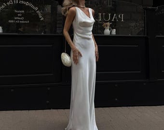 May include: A white, floor-length satin dress with a cowl neckline. The dress is sleeveless and has a simple, elegant design. A small, white clutch is carried on a chain. The background features a dark facade with the word "PAUL" visible.