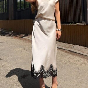 May include: A two-piece set consisting of a short-sleeved, V-neck top and a matching midi-length skirt in a smooth, cream-colored satin fabric. The skirt features a scalloped hemline finished with intricate black lace trim along the bottom edge.