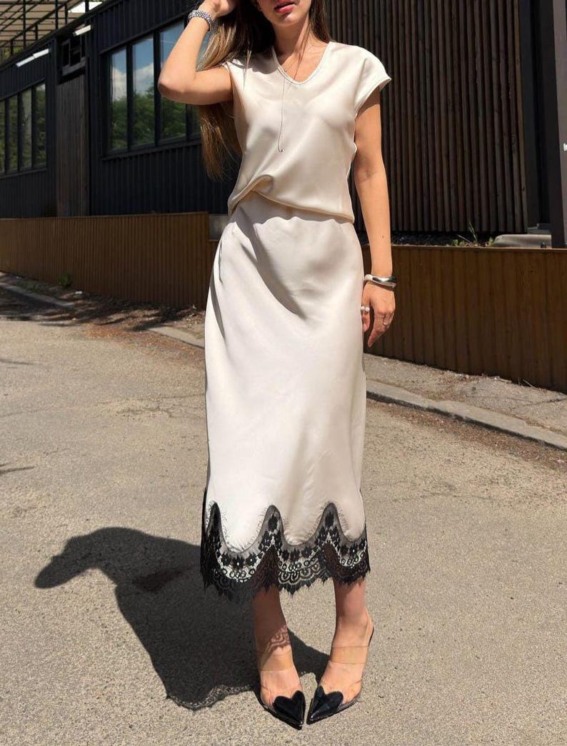 May include: A two-piece set consisting of a short-sleeved, V-neck top and a matching midi-length skirt in a smooth, cream-colored satin fabric. The skirt features a scalloped hemline finished with intricate black lace trim along the bottom edge.