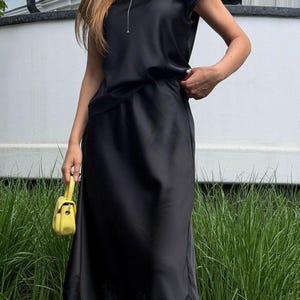 May include: A black satin midi dress featuring a round neckline, short sleeves, and a delicate black lace trim along the hemline. The dress is paired with a small, bright yellow structured handbag with a top handle and a gold-toned clasp.