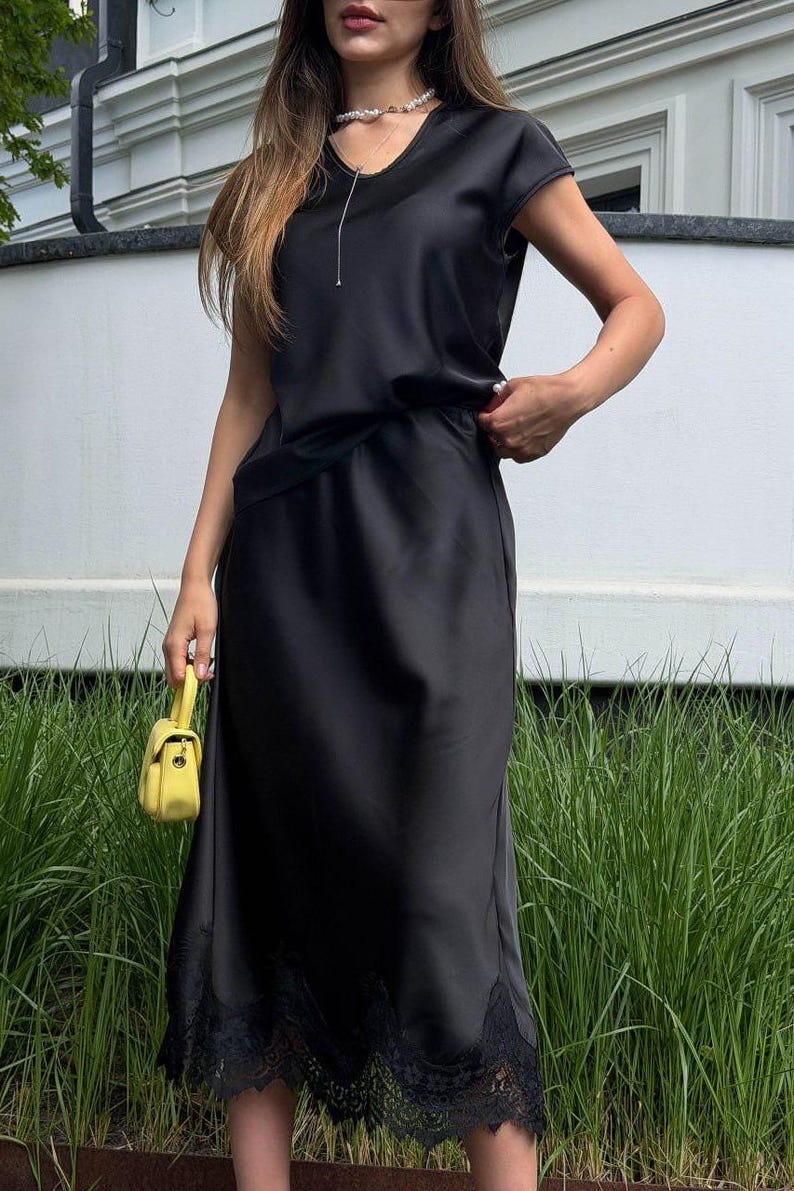 May include: A black satin midi dress featuring a round neckline, short sleeves, and a delicate black lace trim along the hemline. The dress is paired with a small, bright yellow structured handbag with a top handle and a gold-toned clasp.