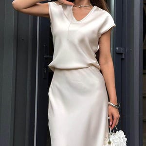 May include: A cream-colored satin two-piece set consisting of a short-sleeved top with a V-neckline and a matching midi-length skirt. The fabric has a smooth, reflective finish. A small white floral-shaped handbag is held in the lower frame.
