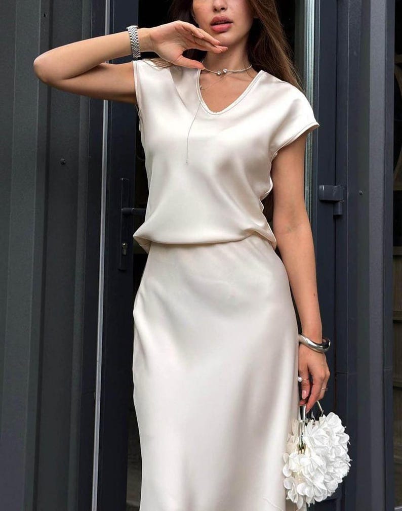 May include: A cream-colored satin two-piece set consisting of a short-sleeved top with a V-neckline and a matching midi-length skirt. The fabric has a smooth, reflective finish. A small white floral-shaped handbag is held in the lower frame.