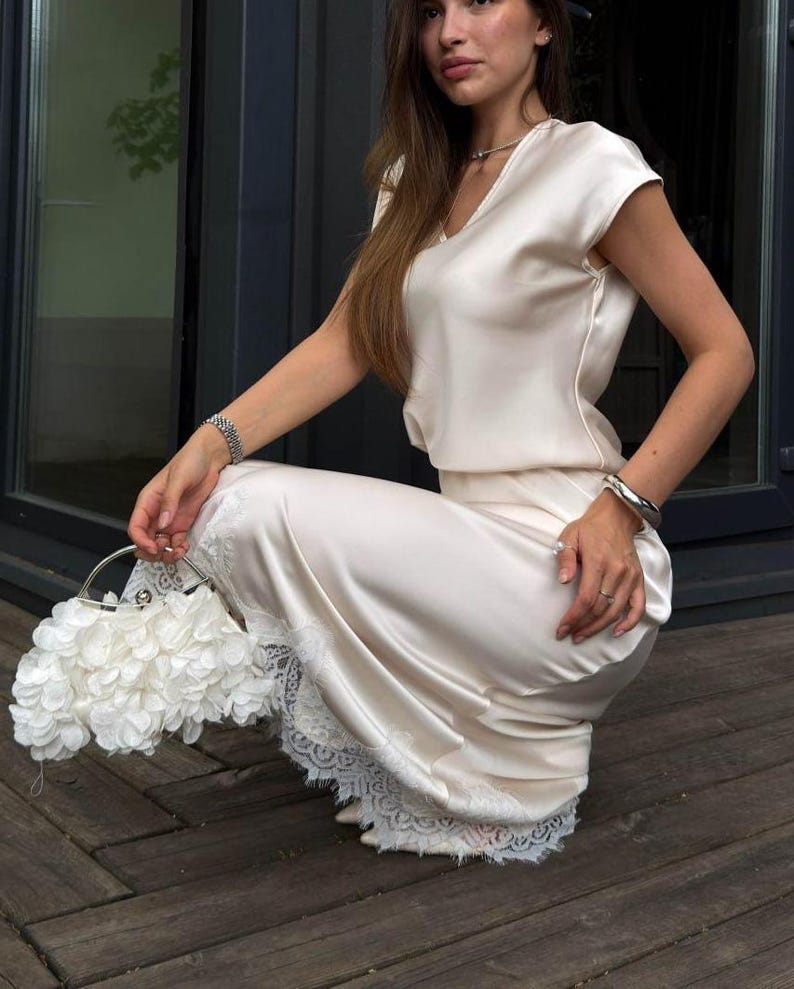 May include: A cream-colored satin two-piece set consisting of a short-sleeved V-neck top and a matching midi skirt with white lace trim at the hem. The set is paired with a white handbag covered in layered fabric flower petals and a silver handle.
