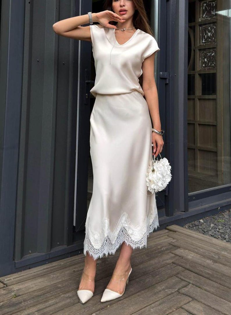 May include: A champagne-colored satin two-piece set consisting of a short-sleeved top with a V-neckline and a matching midi-length skirt. The skirt features a wide, intricate white lace trim along the hemline, creating a layered and textured appearance.