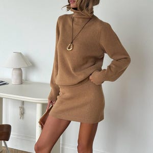Peut inclure: Une robe-pull à manches longues de couleur beige avec un col roulé haut et une texture en maille côtelée. Le vêtement est accompagné d'un long collier en cordon marron foncé portant un grand pendentif ovale lisse de couleur dorée.
