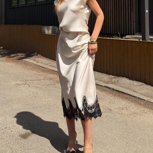 May include: A cream-colored midi dress with a V-neckline and short sleeves, featuring a smooth satin-like finish. The hem of the skirt is adorned with a wide, intricate black lace trim that has a scalloped edge, creating a distinct contrast.