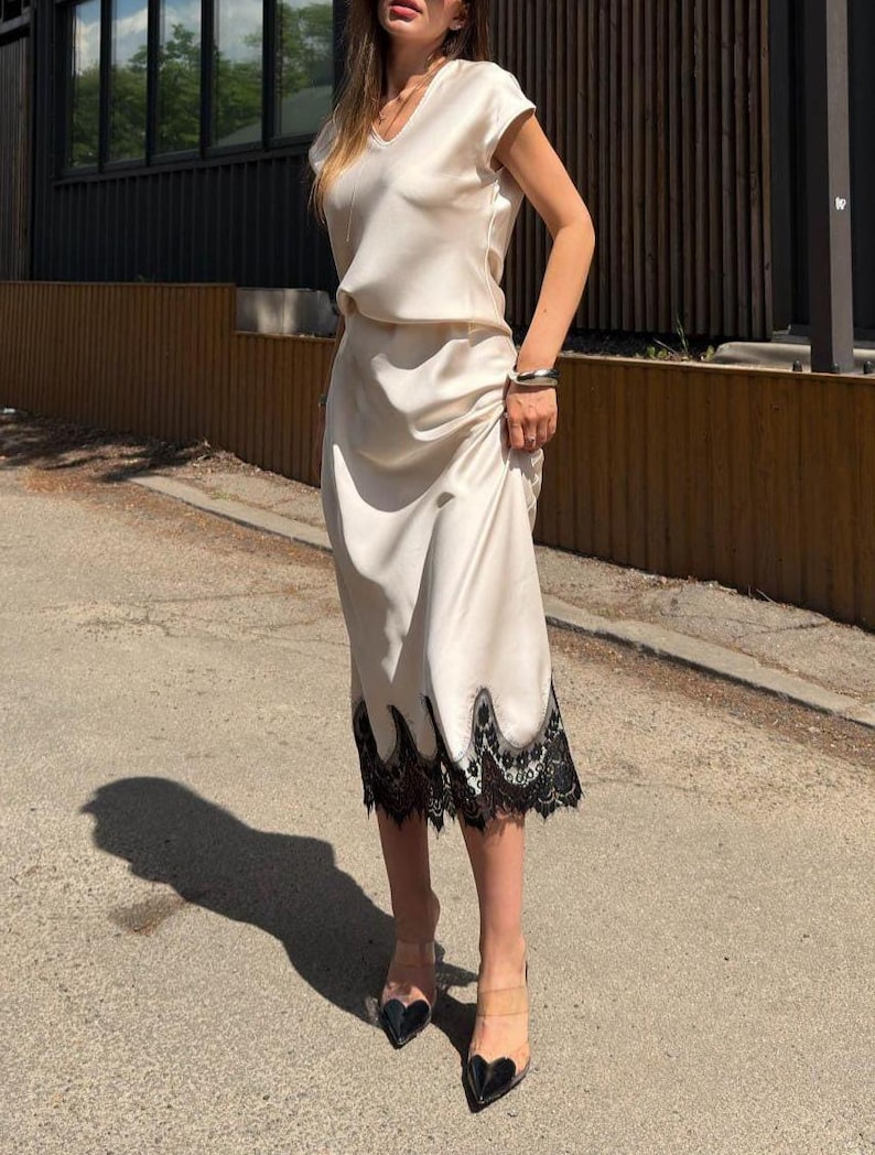 May include: A cream-colored midi dress with a V-neckline and short sleeves, featuring a smooth satin-like finish. The hem of the skirt is adorned with a wide, intricate black lace trim that has a scalloped edge, creating a distinct contrast.
